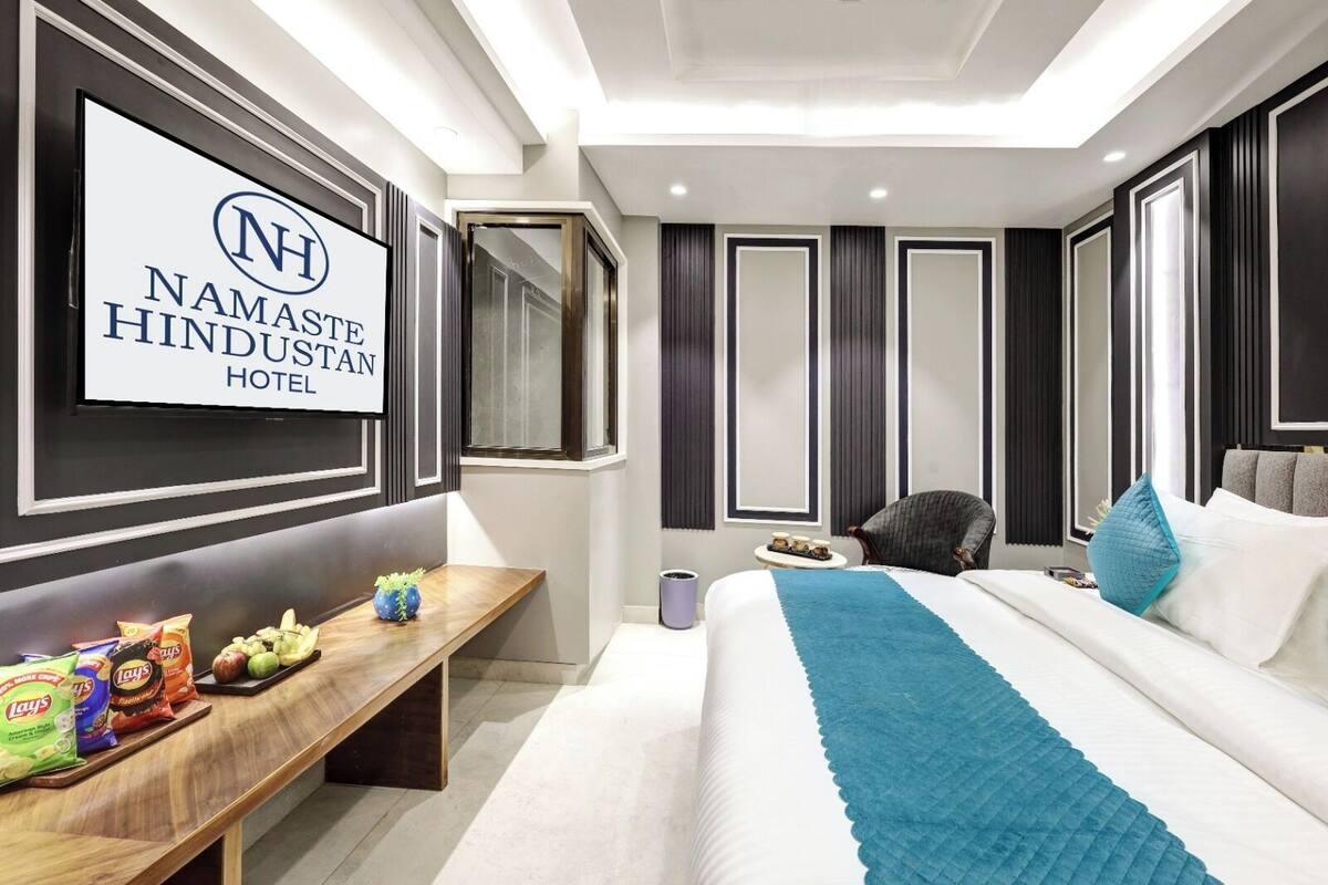 Quarto de hotel com uma cama, uma escrivaninha com lanches, uma cadeira e uma televisão exibindo o nome do hotel.