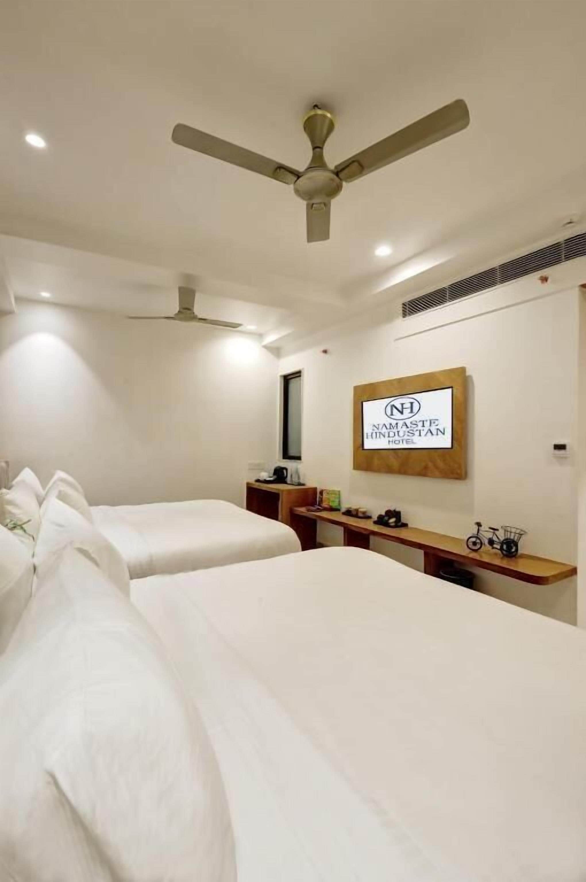 Habitación de hotel con dos camas, un ventilador de techo, un televisor montado en la pared y un cartel del Hotel Namaste Hindustan.