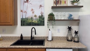 A kitchen with a granite countertop, a black sink, a Samsung dishwasher, a painting of palm trees, and decorative items on shelves.