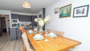 Un coin repas avec une table en bois dressée pour quatre personnes, un vase de fleurs blanches et une cuisine en arrière-plan.