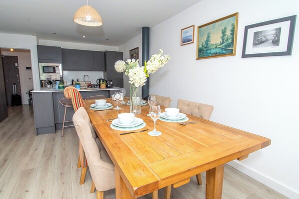 Un comedor con una mesa de madera dispuesta para cuatro personas, un jarrón de flores blancas y una cocina al fondo.