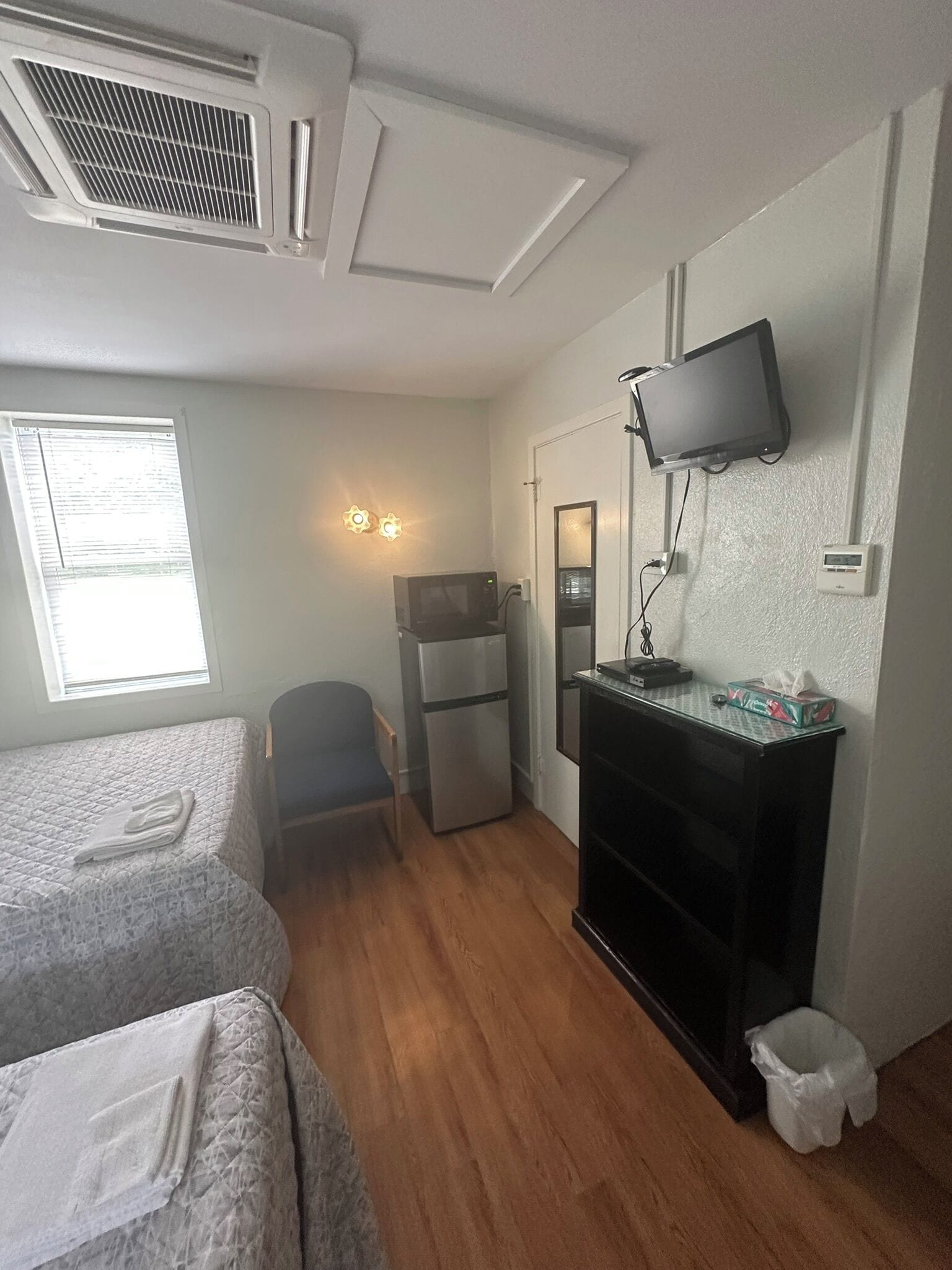 A compact hotel room with two beds, a small kitchenette, a TV mounted on the wall, and a window with blinds.
