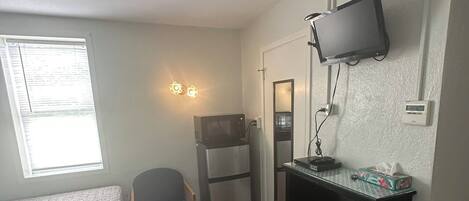 A compact hotel room with two beds, a small kitchenette, a TV mounted on the wall, and a window with blinds.