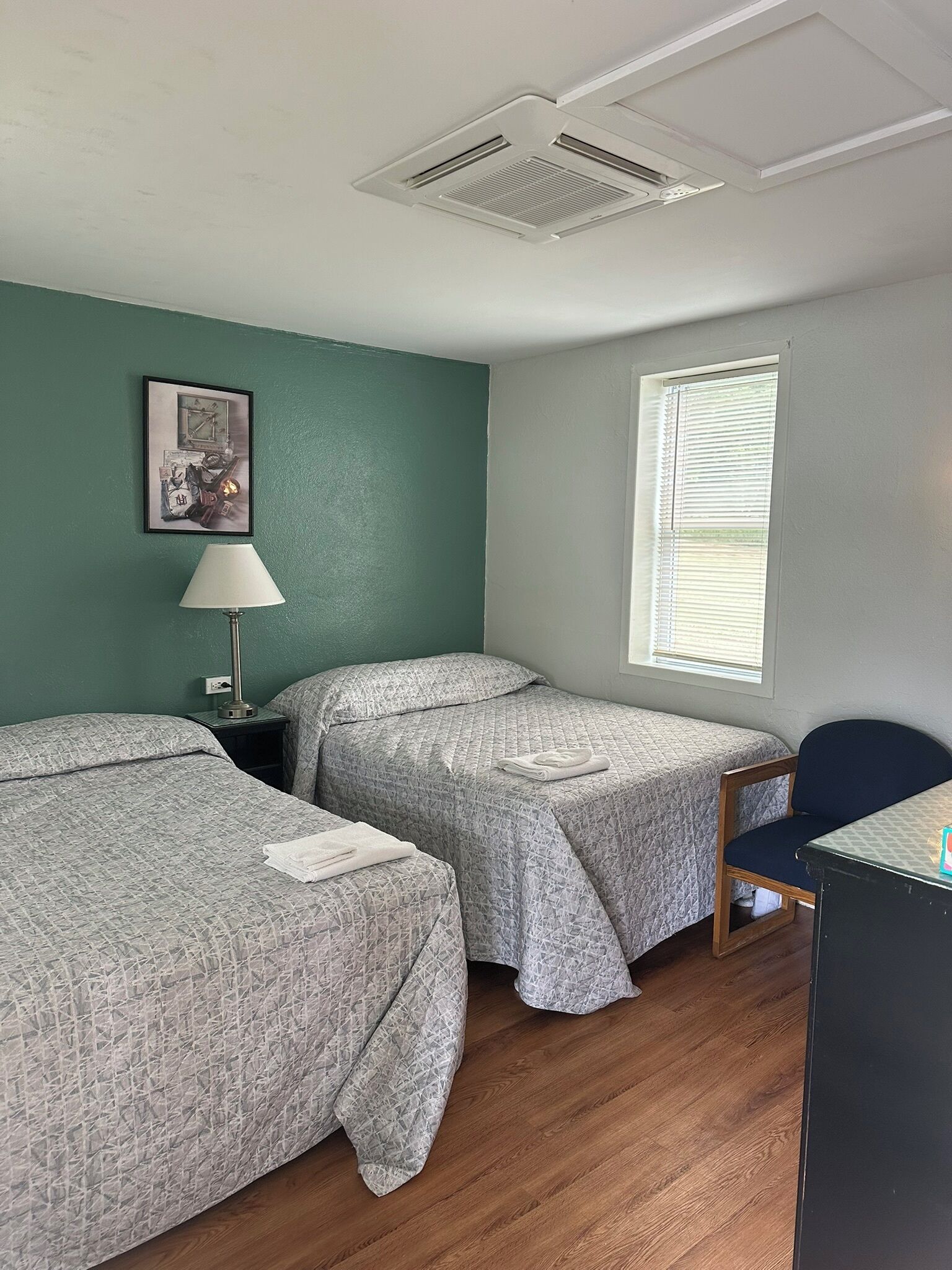 A room with two beds, a desk, a chair, a window with blinds, a picture on the wall, and an air vent.