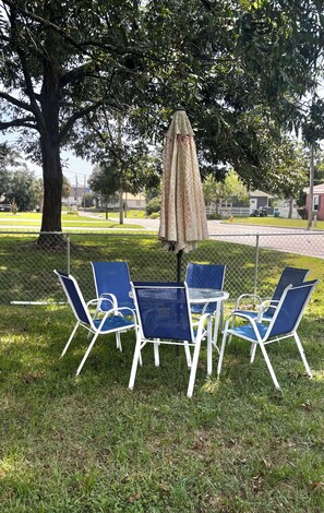 A picnic setup with a table and four chairs under a patterned umbrella, surrounded by a grassy area and trees. - Spacious family beach home wgrill,arcade, pool table,pingpong, gameroom,fire pit (Gulfport)