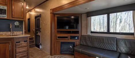 Interior compacto de autocaravana con kitchenette, zona de estar y un ventanal grande con vistas a árboles.
