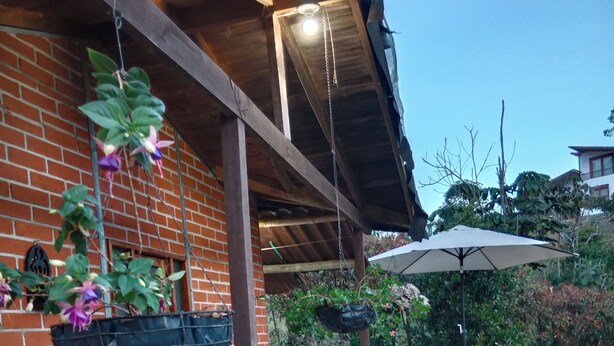 Eine überdachte Terrasse mit einer Backsteinmauer, einer blauen Bank mit Kissen, hängenden Pflanzgefäßen, einem Tisch mit Stühlen und einem Sonnenschirm.