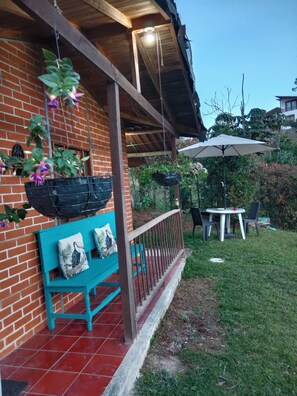 A covered patio with a brick wall, a blue bench with cushions, hanging planters, a table with chairs, and an umbrella.