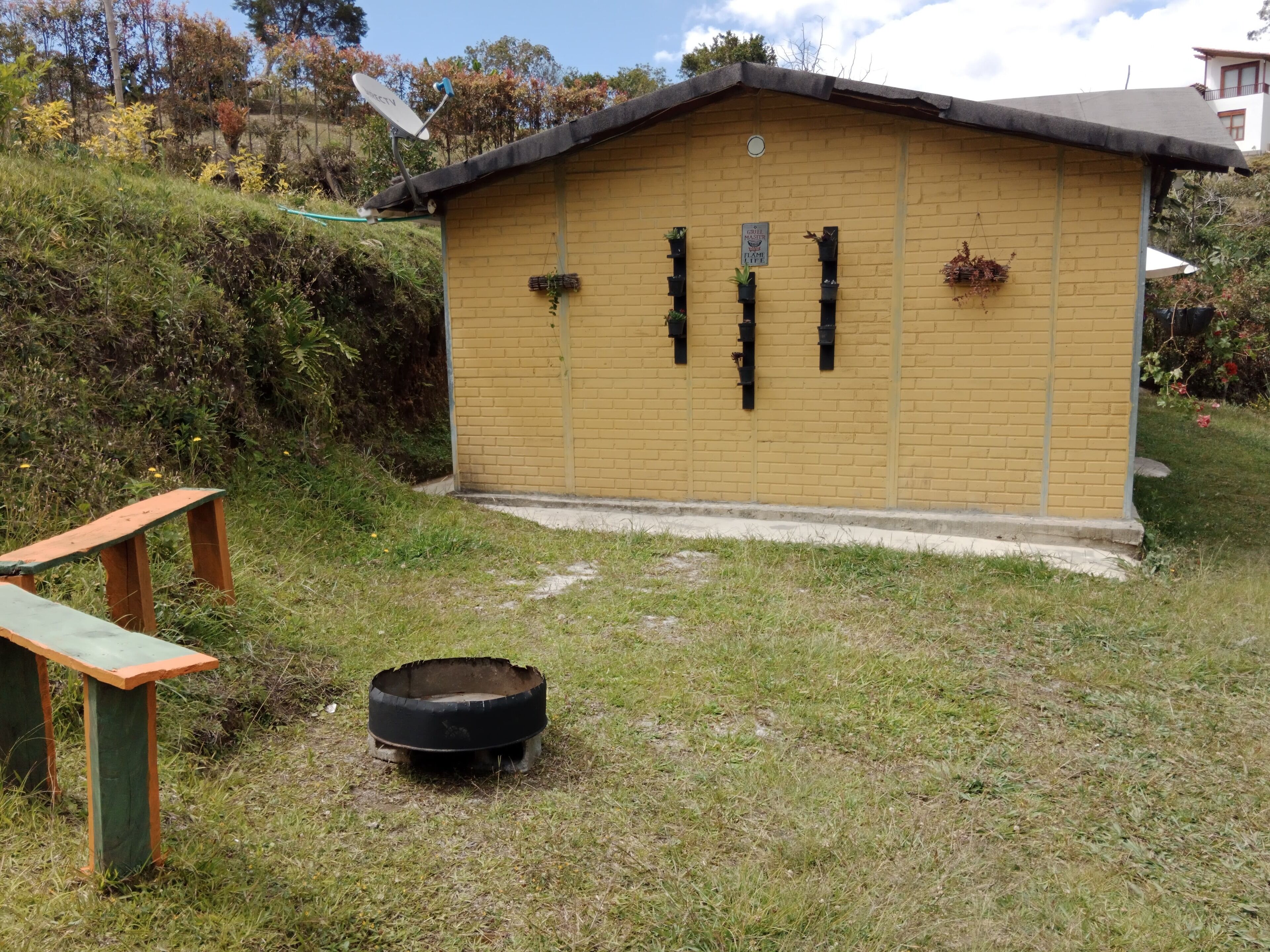 A small, yellow brick building with a flat roof, surrounded by grass and bushes. There is a wooden bench and a tire in the foreground.