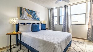 Habitación de hotel con una cama grande, un ventilador de techo, un cuadro sobre la cama y una ventana con cortinas.