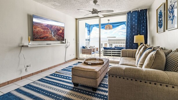 Una sala de estar con un televisor grande, un ventilador de techo, un sofá y una alfombra estampada.