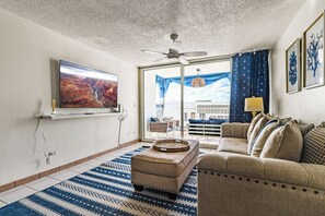 Ein Wohnzimmer mit einem großen Fernseher, einem Deckenventilator, einer Couch und einem gemusterten Teppich.