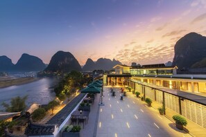 A riverside terrace with outdoor seating, a modern building, and mountainous views. - YangShuo Percent Hotel (Guilin)