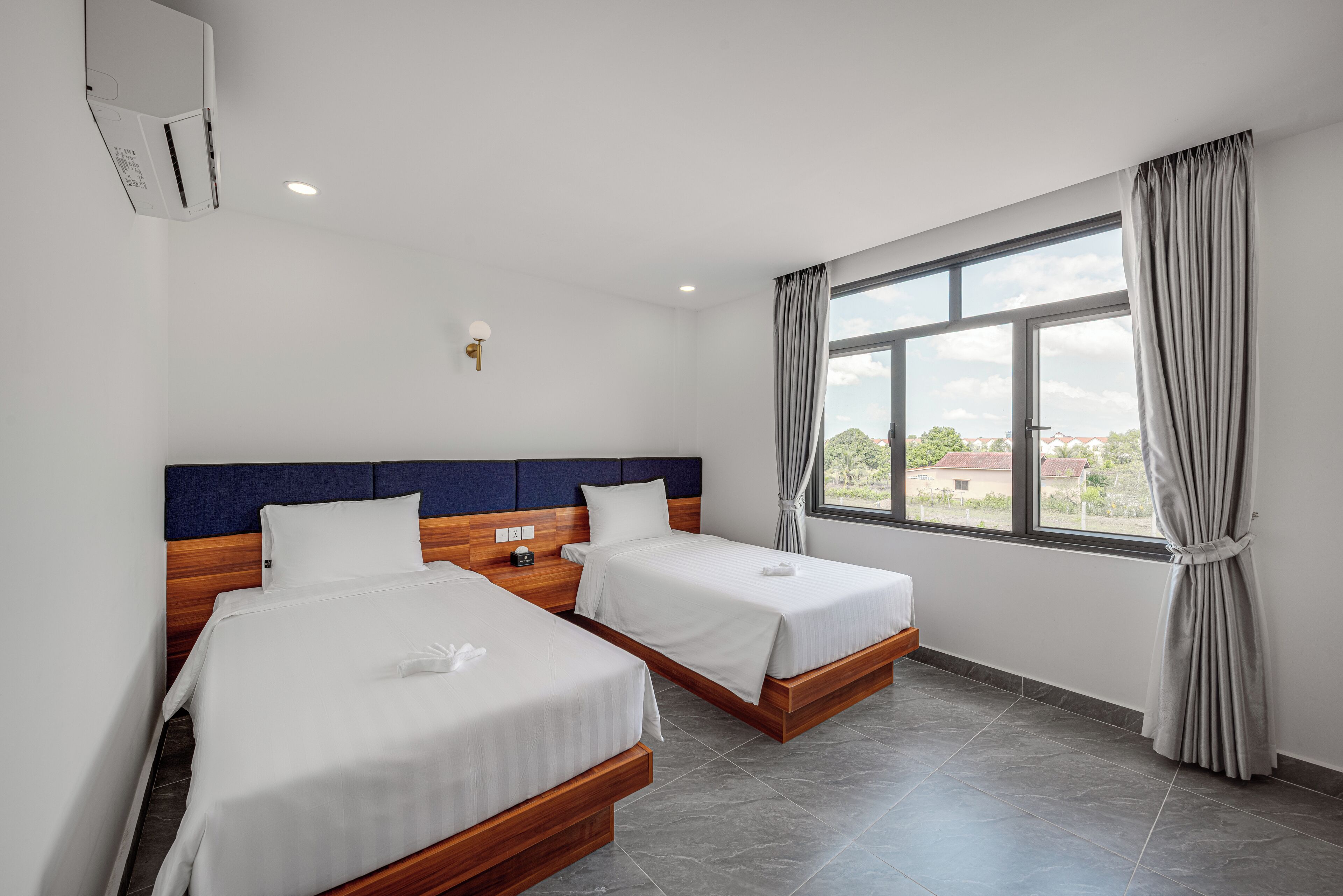 Ett hotellrum med två sängar, ett stort fönster med gardiner, en sänggavel i trä och en väggmonterad luftkonditioneringsanläggning.