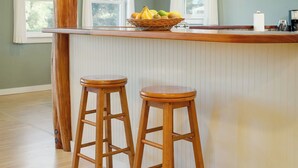 A kitchen with a wooden island, two wooden stools, a fruit bowl, and a window with curtains. - Sunset Orchard- Sunny side of Waimea, close to beaches! (Waimea)