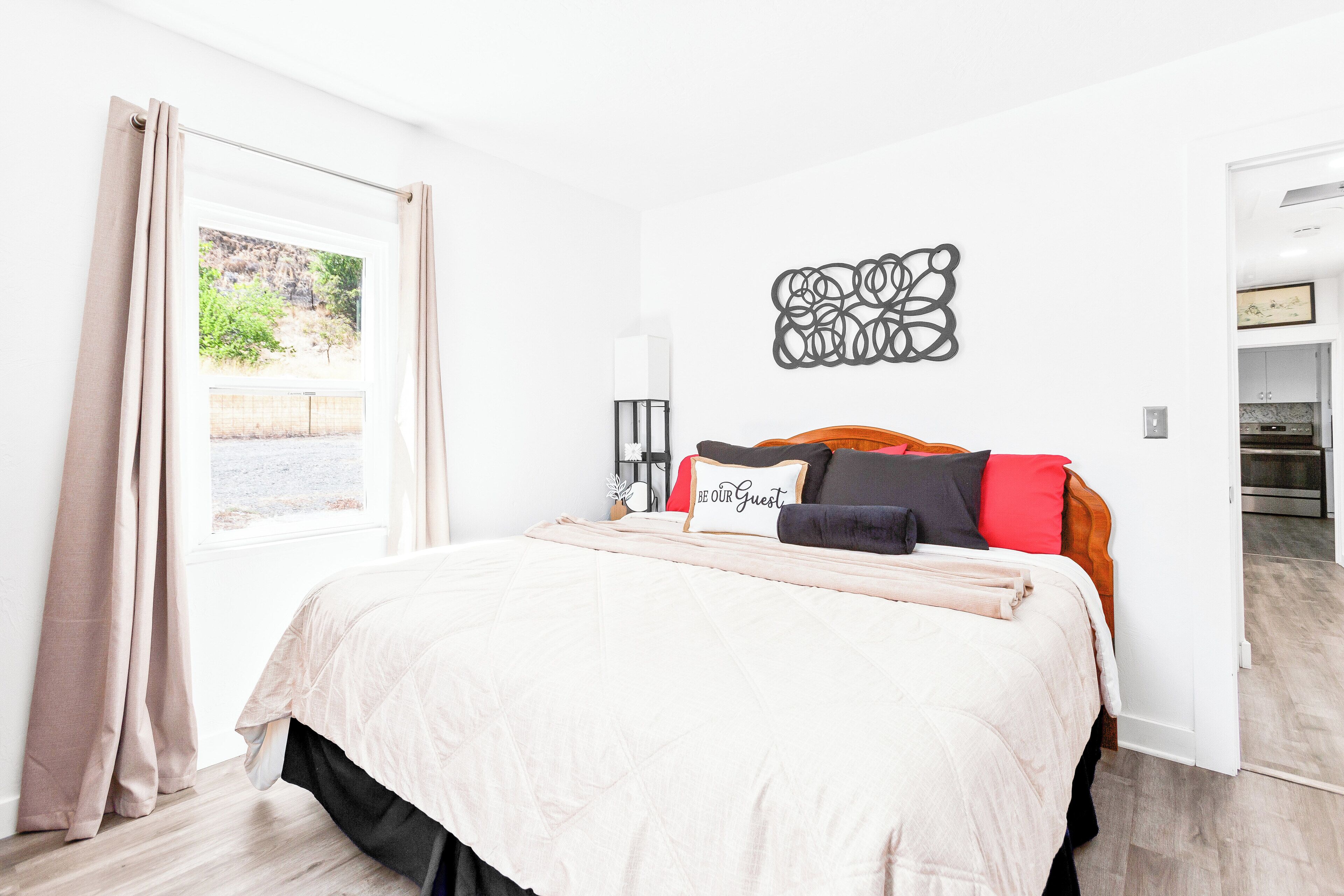 Un dormitorio con cama, una ventana con cortinas, una obra de arte en la pared y vista a una cocina.