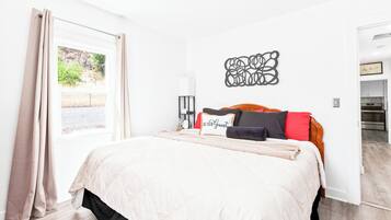 Un dormitorio con cama, una ventana con cortinas, una obra de arte en la pared y vista a una cocina.