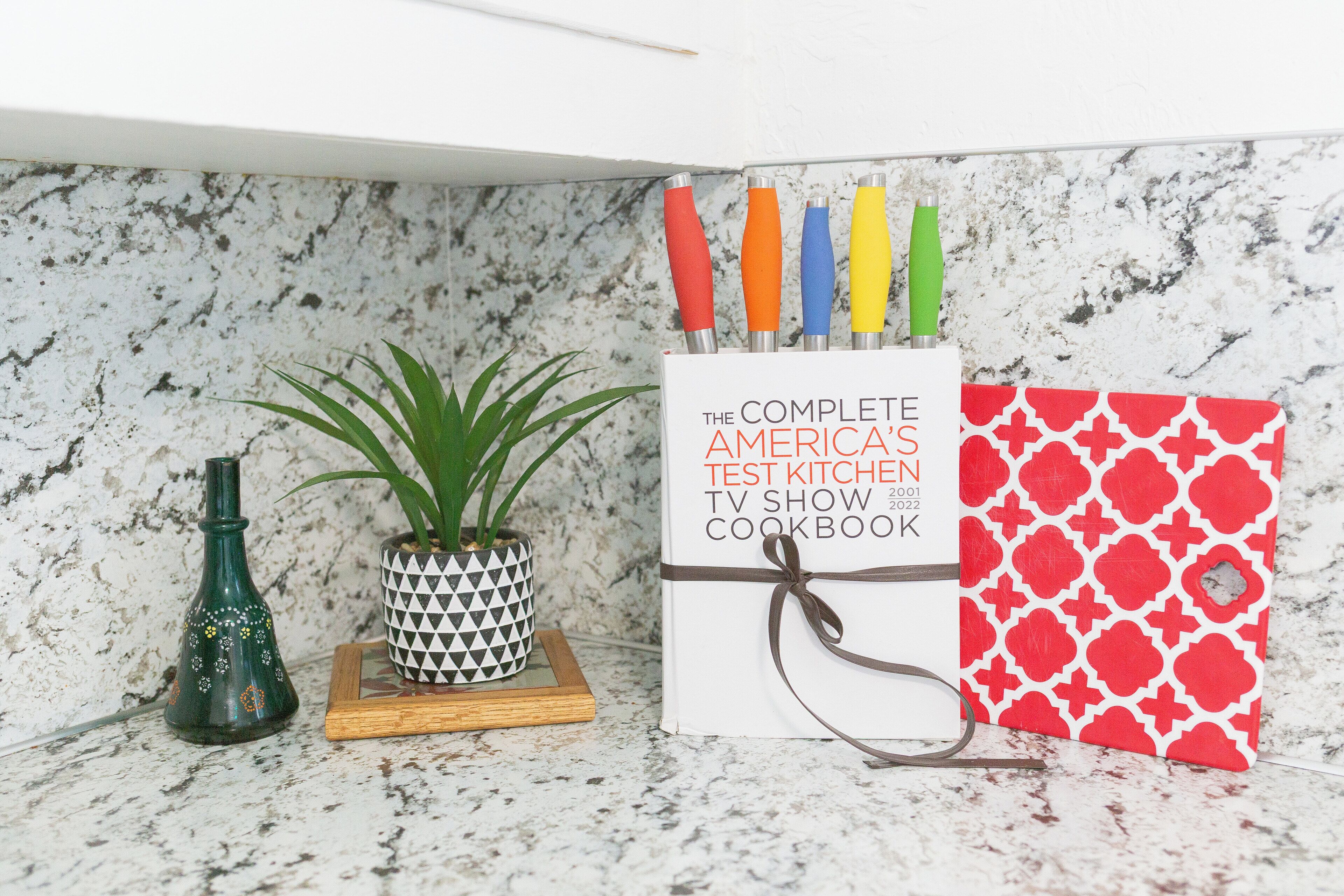 Un plan de travail de cuisine avec un livre de recettes intitulé « Le livre de recettes complet de l’émission de télévision America’s Test Kitchen », une plante en pot et une bouteille décorative.