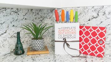 Un plan de travail de cuisine avec un livre de recettes intitulé « Le livre de recettes complet de l’émission de télévision America’s Test Kitchen », une plante en pot et une bouteille décorative.