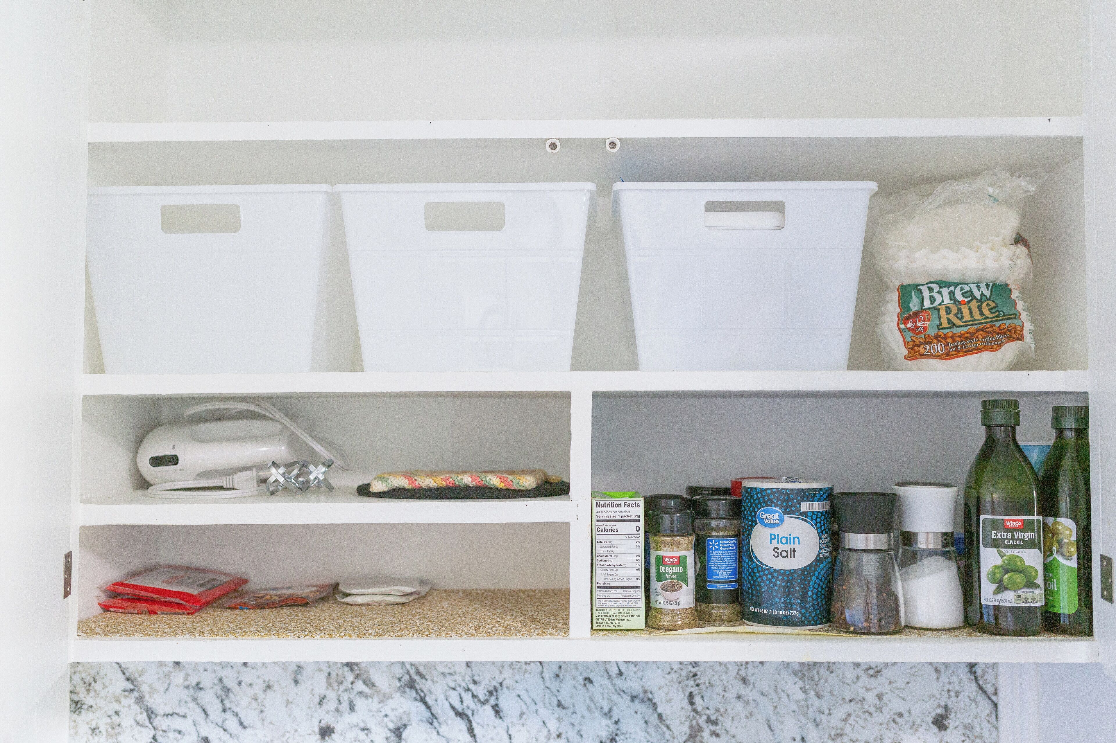 Una cocina con armarios blancos, una cafetera y diversos alimentos, incluyendo un paquete de café y botellas de aceite de oliva.