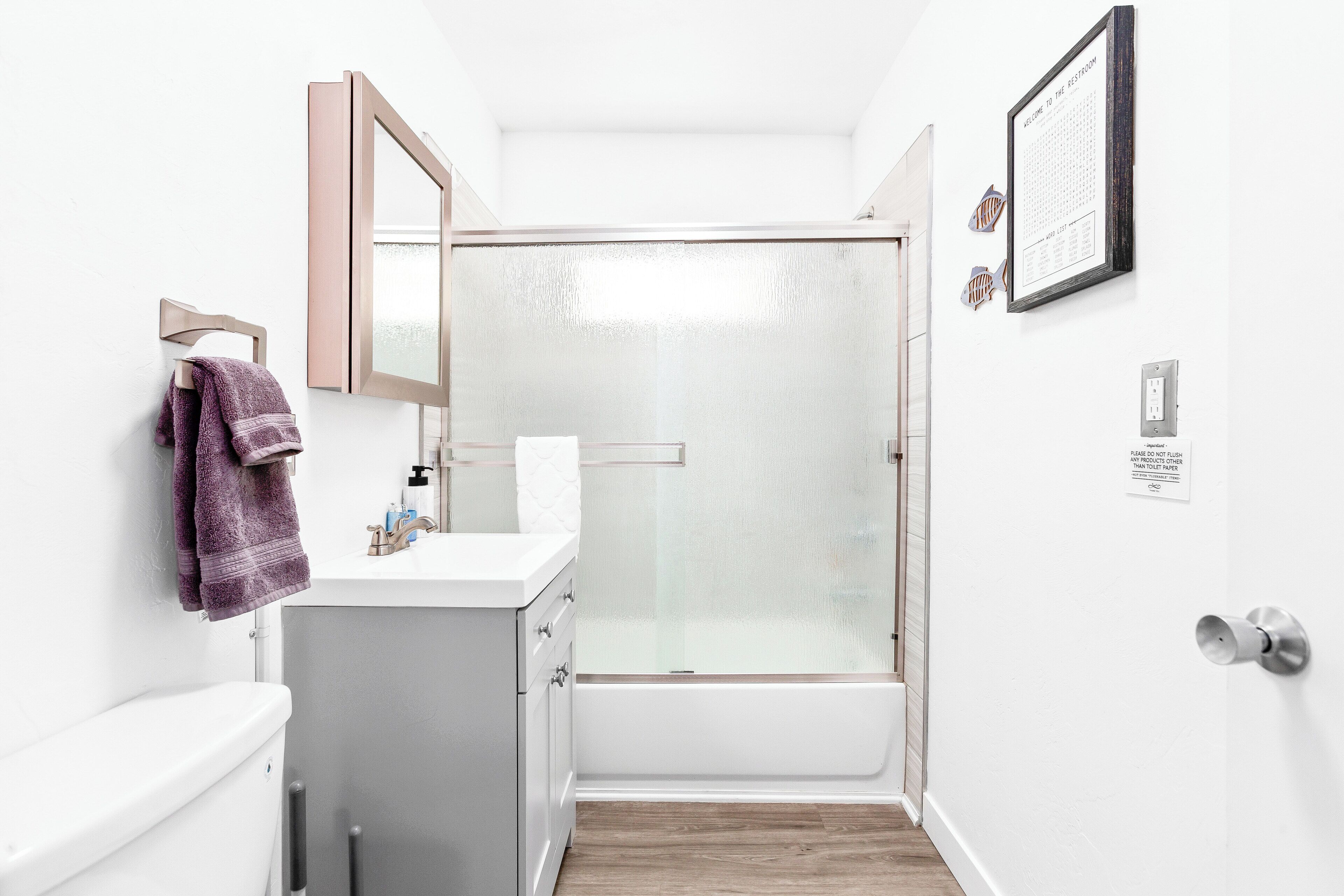Une petite salle de bain blanche, avec une douche, des toilettes et un lavabo. Une serviette est accrochée au mur, et une liste de mots encadrée est également fixée au mur.