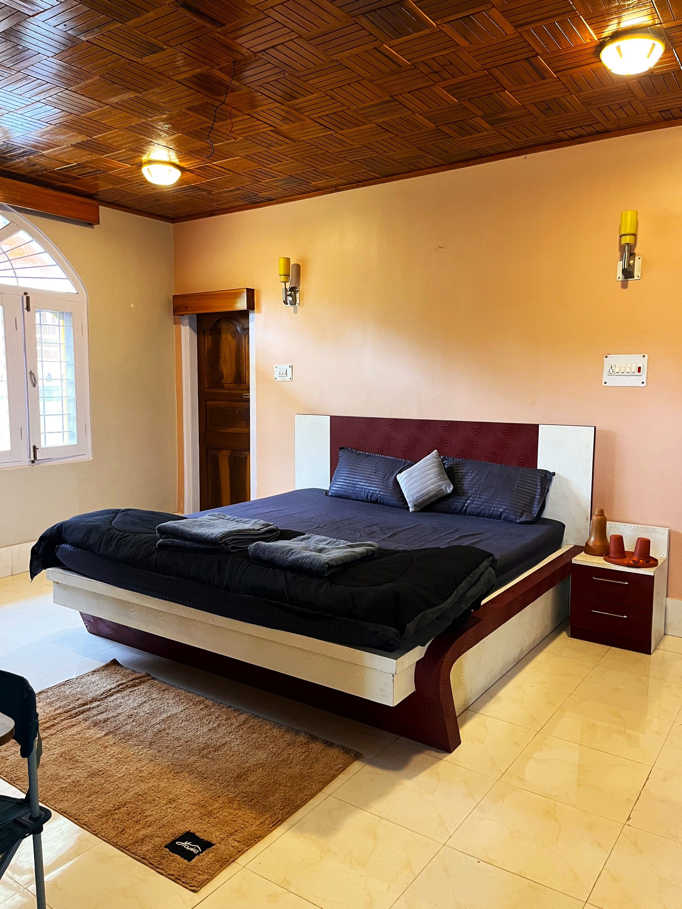 A bedroom with a wooden ceiling, a bed with a dark headboard, a nightstand, and a small table with two cups.