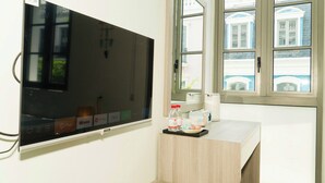 Ett hotellrum med en stor platt-TV som är monterad på väggen, ett litet skrivbord i trä med ett bricka som innehåller en flaska, en kopp och en näsdukask.
