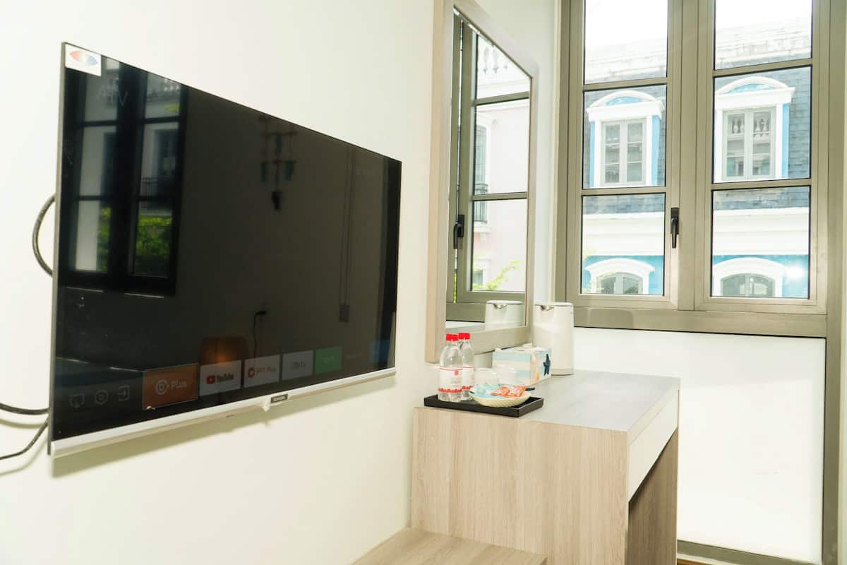 Habitación de hotel con un televisor de pantalla plana grande montado en la pared, un pequeño escritorio de madera con una bandeja que contiene una botella, una taza y una caja de pañuelos.