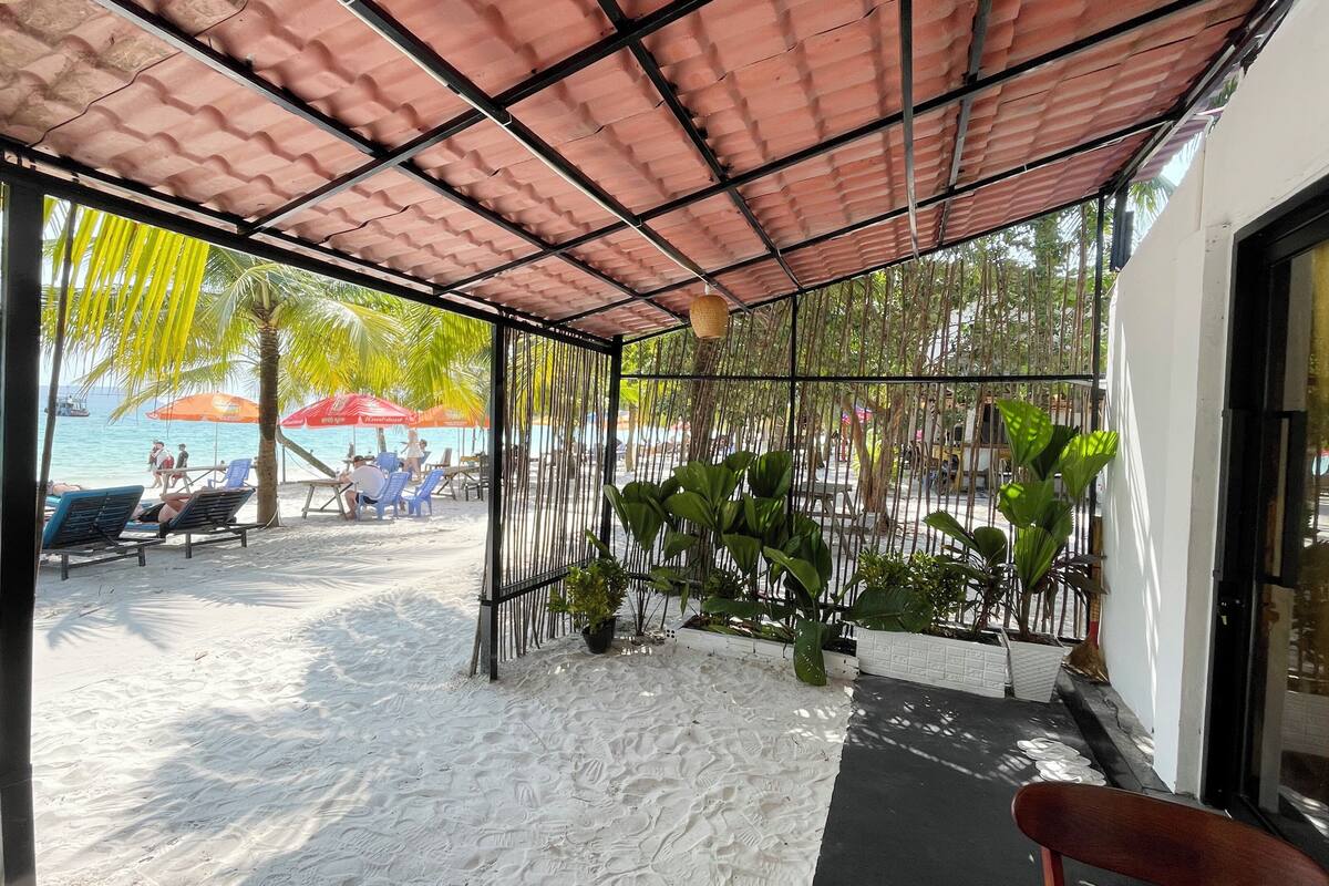 A covered outdoor area with a sandy floor, blue lounge chairs, and potted plants.