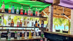 Een bar met meerdere flessen drank, een televisie en uitzicht op palmbomen en een strand.