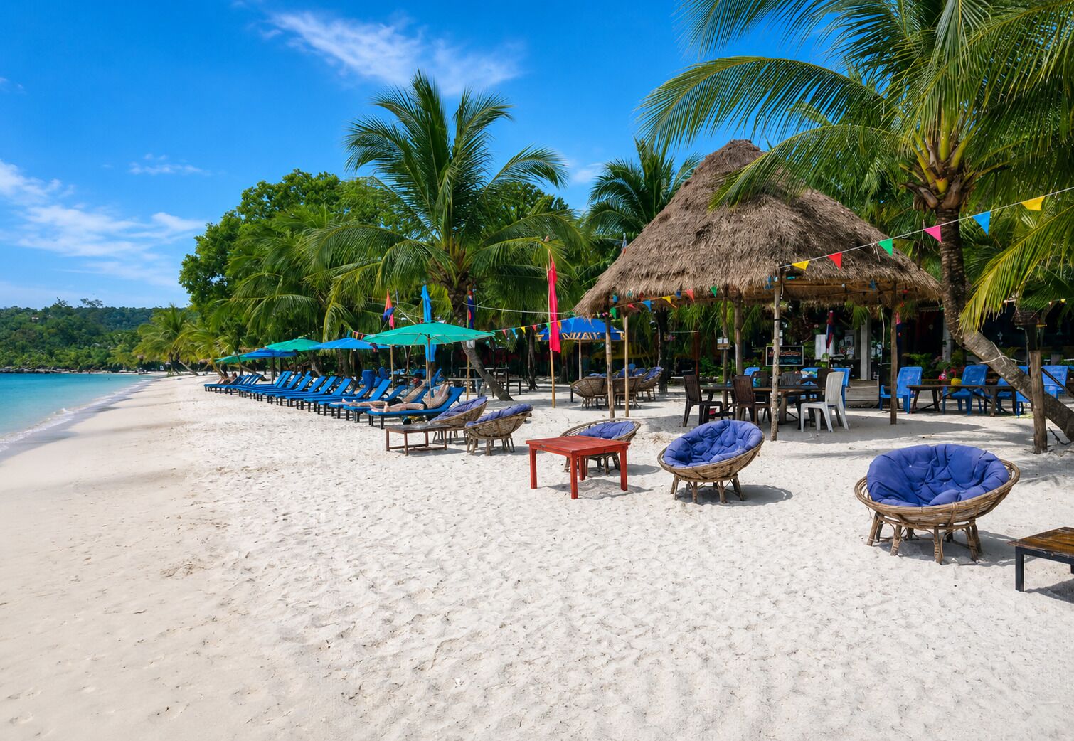 Une plage avec des chaises longues et des tables placées sous des parasols en feuilles de palmier, des palmiers et un ciel bleu dégagé.