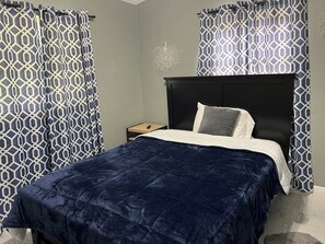 Ein ordentlich bezogenes Bett mit einer dunkelblauen Tagesdecke, einem weißen und einem grauen Kopfkissen auf einem schwarzen Kopfteil. Ein kleiner Nachttisch aus Holz mit einer Fernbedienung steht daneben. Das Zimmer hat bunte Vorhänge und eine dekorative Wandleuchte.