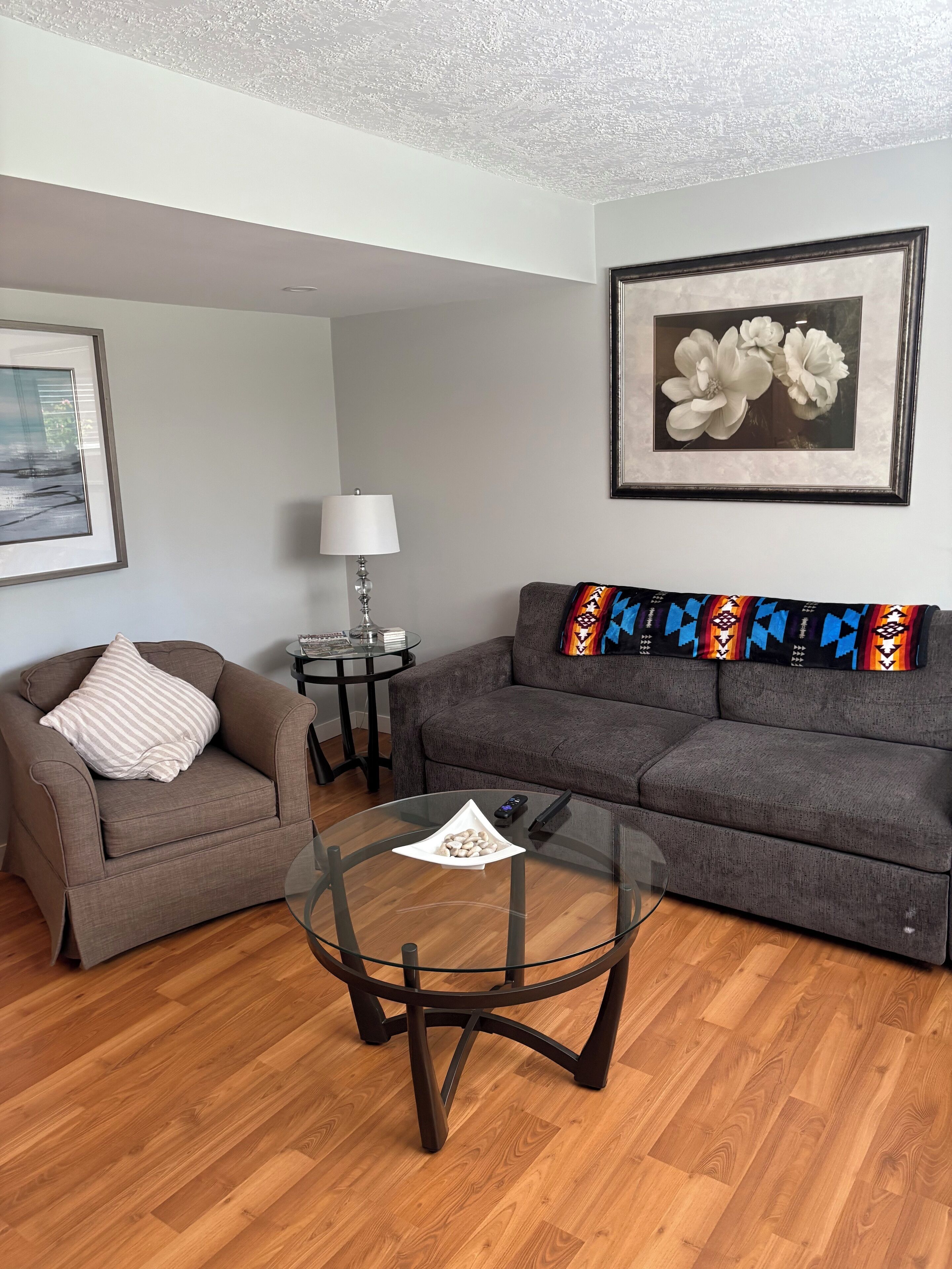 A living room with a grey sofa, a round glass coffee table, a striped armchair, and a framed picture on the wall.