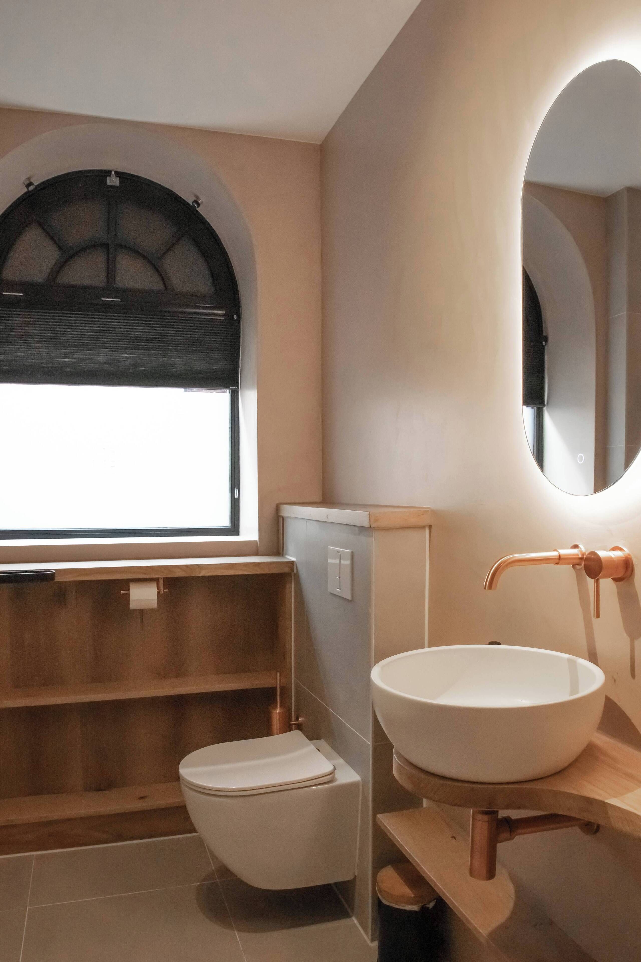 Un baño moderno con inodoro, lavamanos con grifo de cobre y ventana con persiana negra.