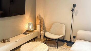 A modern living room with a flat-screen TV mounted on the wall, a round coffee table, a beige sofa, a floor lamp, and a decorative vase with pampas grass.