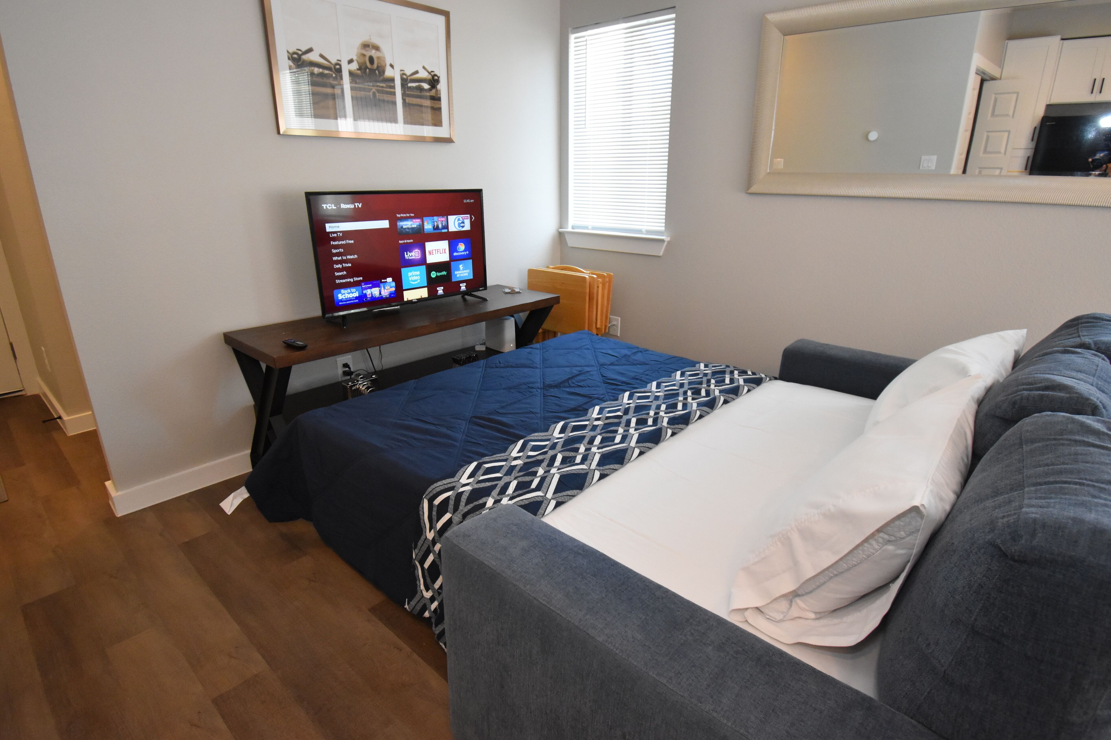 Ein Schlafzimmer mit Bett, Schreibtisch mit Computer, Fenster mit Jalousien und Spiegel an der Wand.