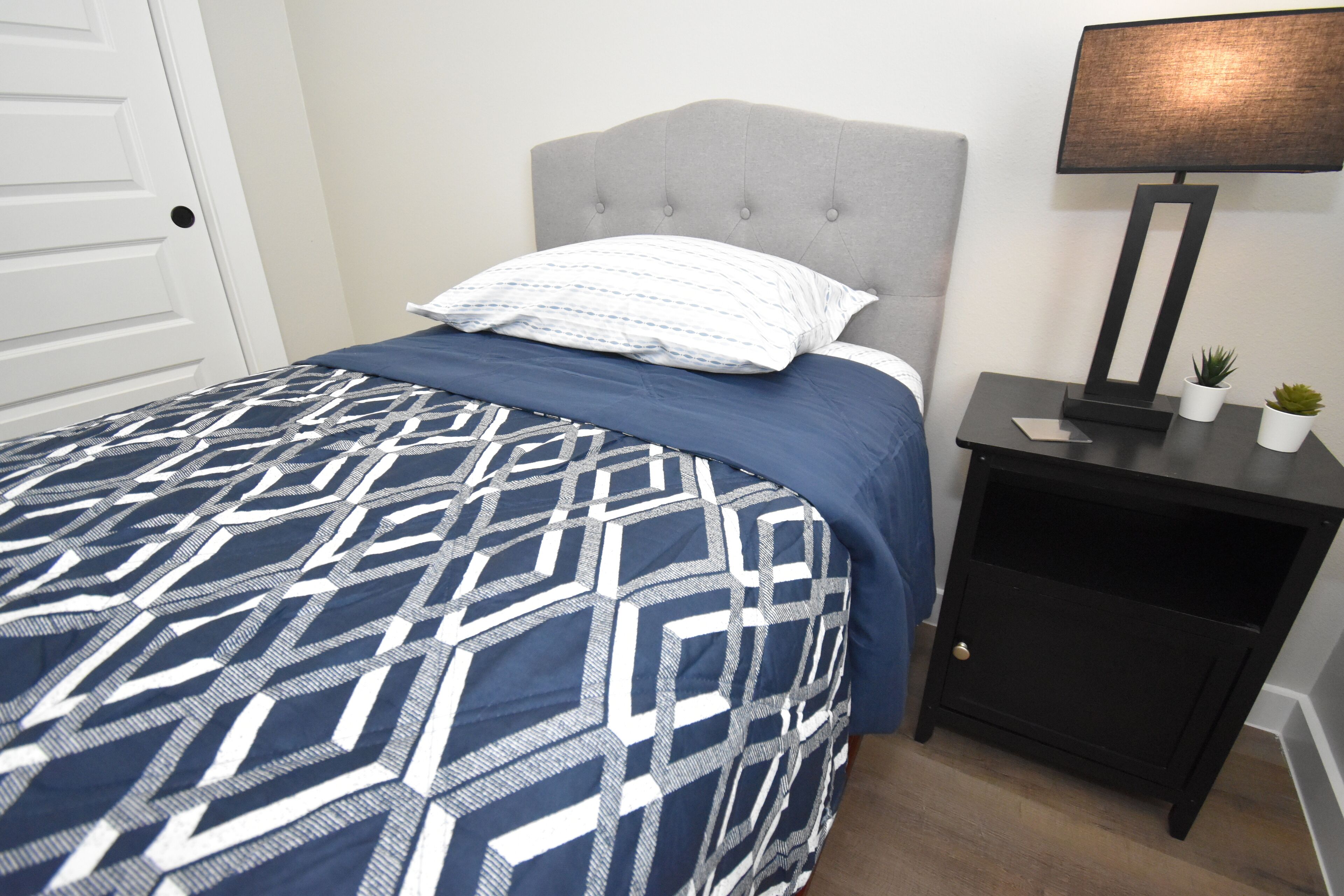 Une chambre avec une couvre-lit à motifs, une tête de lit capitonnée, une table de chevet avec une lampe et une petite plante.