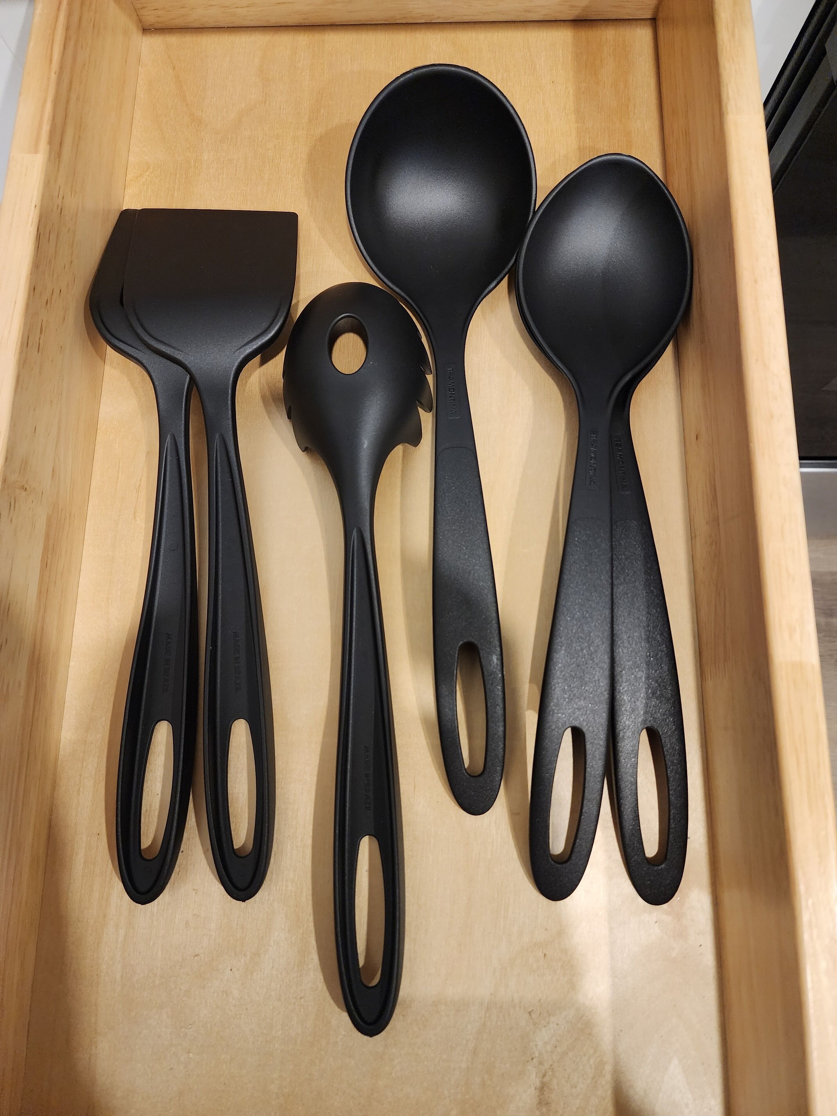A wooden drawer containing four black kitchen utensils: a spatula, a ladle, and two spoons.