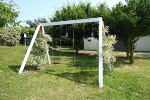 A white swing set in a grassy yard with trees and a building in the background.