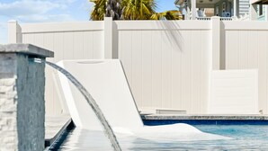 A swimming pool with a waterfall feature, surrounded by a white fence and palm trees. - 5BR Channel Home w/ Heated Pool + Fire Pit (North Myrtle Beach)