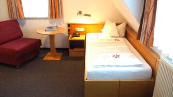A hotel room with a bed, a red armchair, a small table, and a nightstand with a lamp.