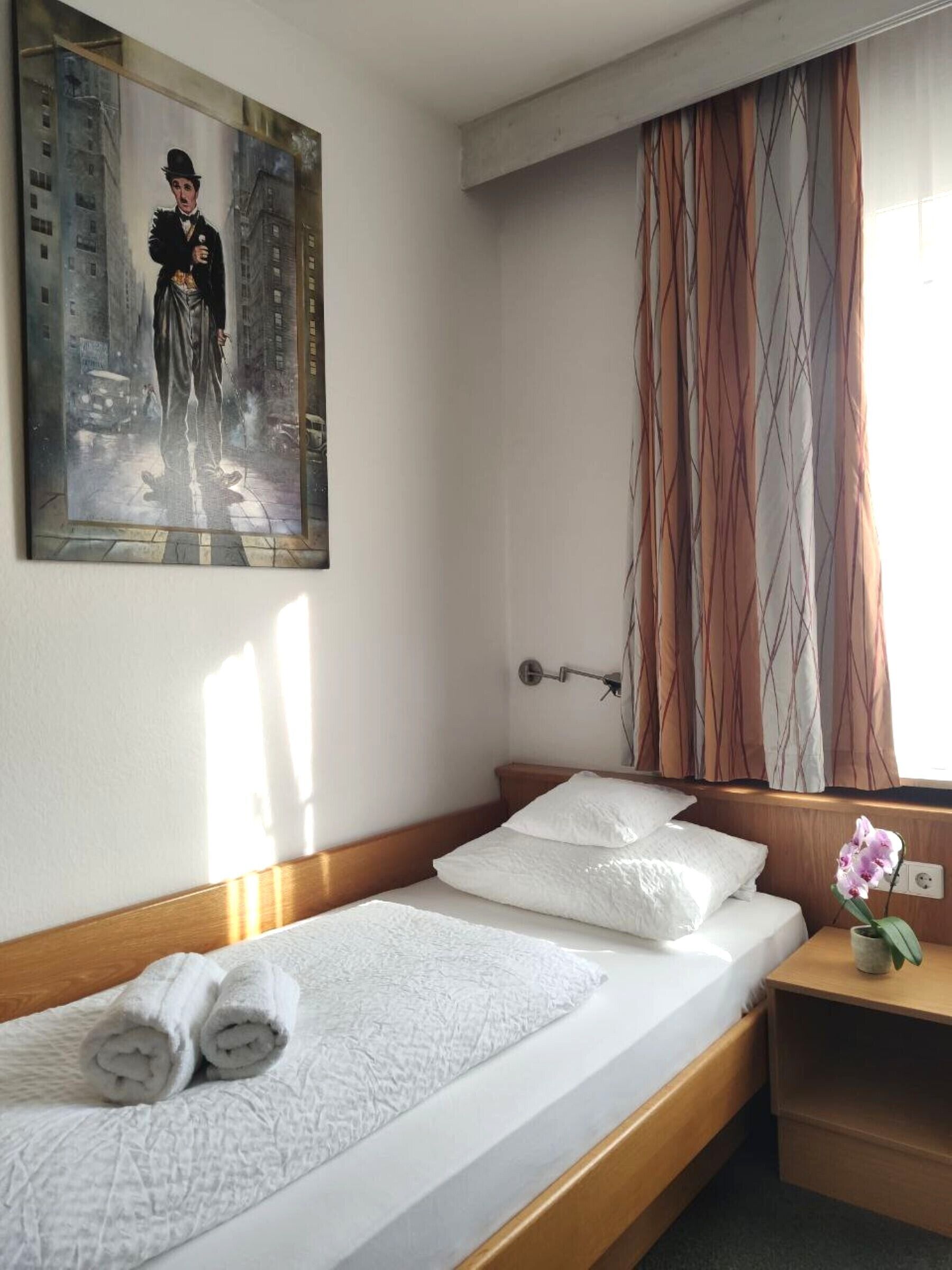Una habitación de hotel con una cama individual, una mesita de noche con un florero y un cuadro en la pared, además de una ventana con cortinas.