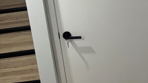 Eine weiße Tür mit Schlüsselloch und einem hineingesteckten Schlüssel, neben einer Holztreppe mit schwarzen Stufen.