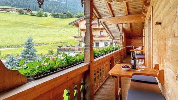 En træterrasse med et bord og siddepladser, med udsigt over et grønt landskab og en bygning med altaner.