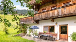 A wooden chalet with a balcony, a stone patio, and a small garden area with a bench and table. - Two big alpine stays for families & groups (Alpbach)