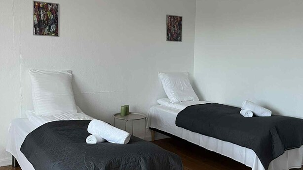 Twee eenpersoonsbedden met witte lakens en donge dekens, een klein rond tafeltje met een groene kaars, en twee abstracte kunstwerken aan de muur.