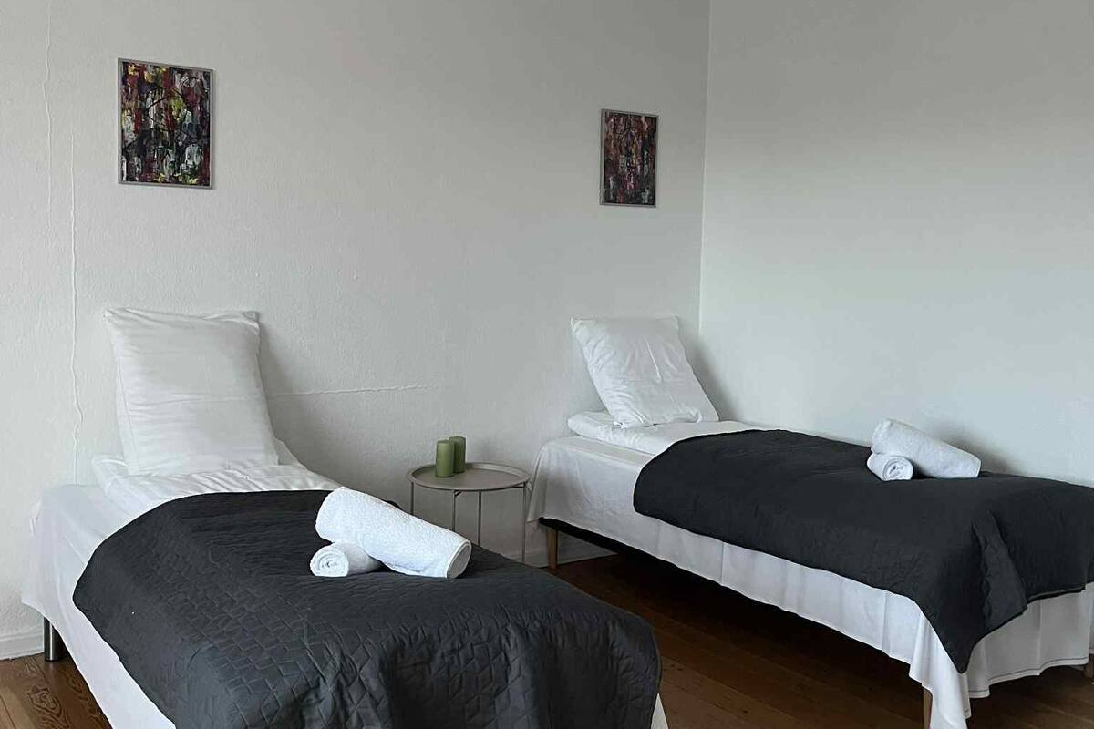Two single beds with white linens and dark quilts, a small round table with a green candle, and two abstract art pieces on the walls.