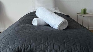 Een net opgemaakt bed met een gewatteerde donkergrijze deken en twee opgerolde witte handdoeken.