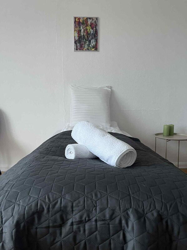 A neatly made bed with a quilted dark grey bedspread and two rolled white towels.