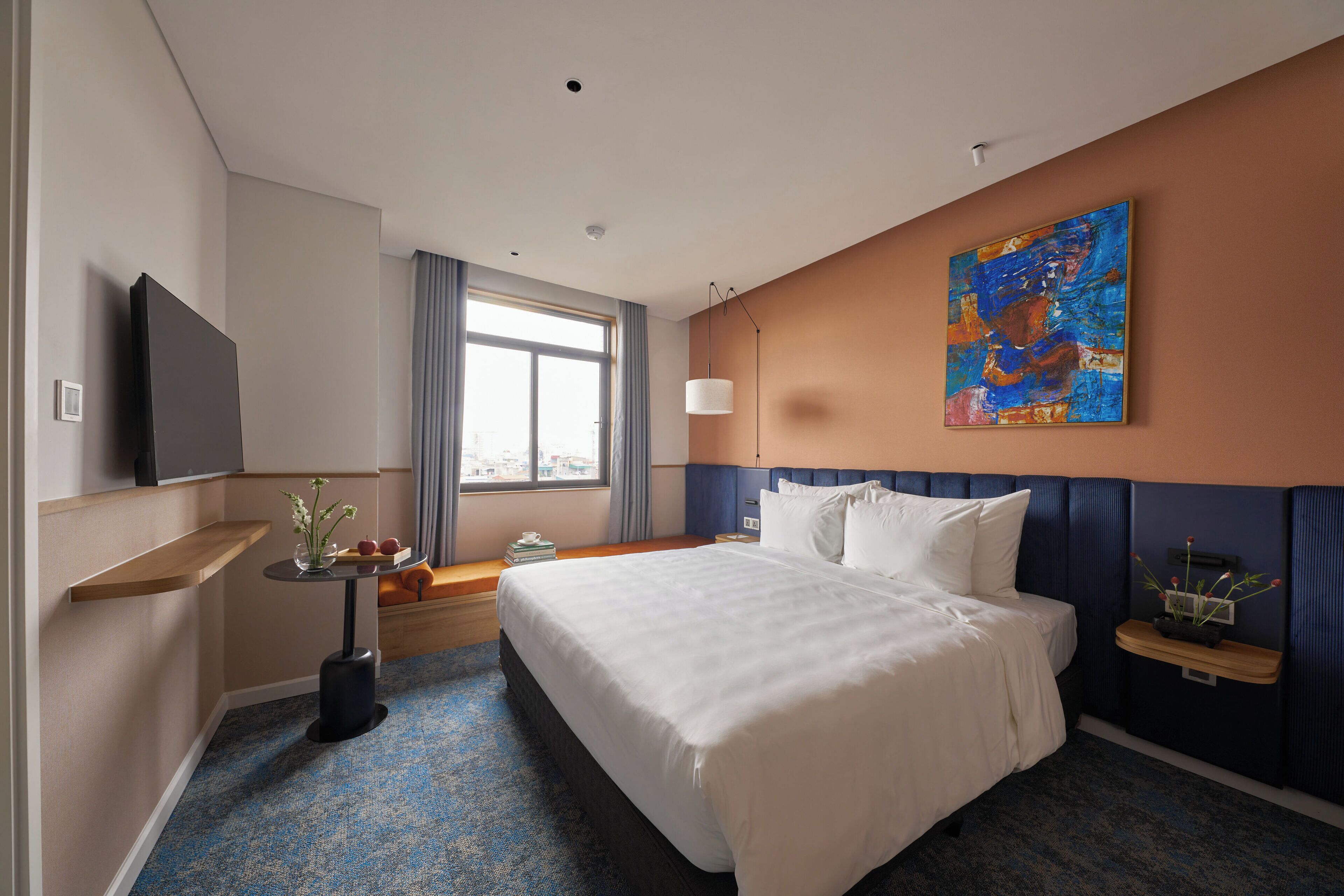 A hotel room with a large bed, a flat-screen TV, a small table with a vase, and a window with curtains.